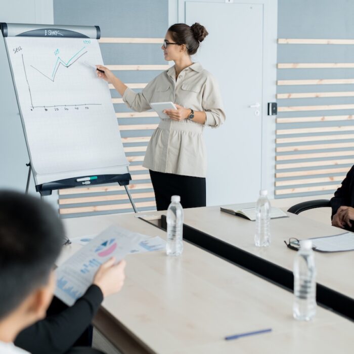 Young sales coach with hair bun wearing glasses holding presentation and explaining graph to business people at training class